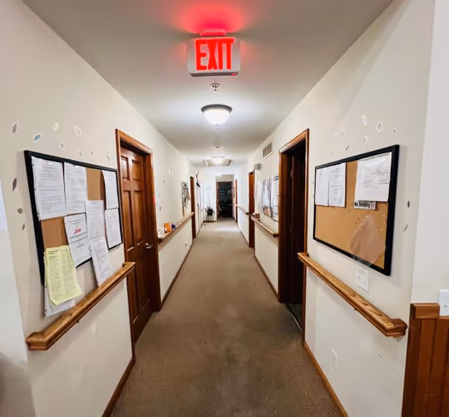 Long carpeted interior hallway in an assisted living facility with bulletin boards, wooden doors, handrails, and an illuminated EXIT sign.