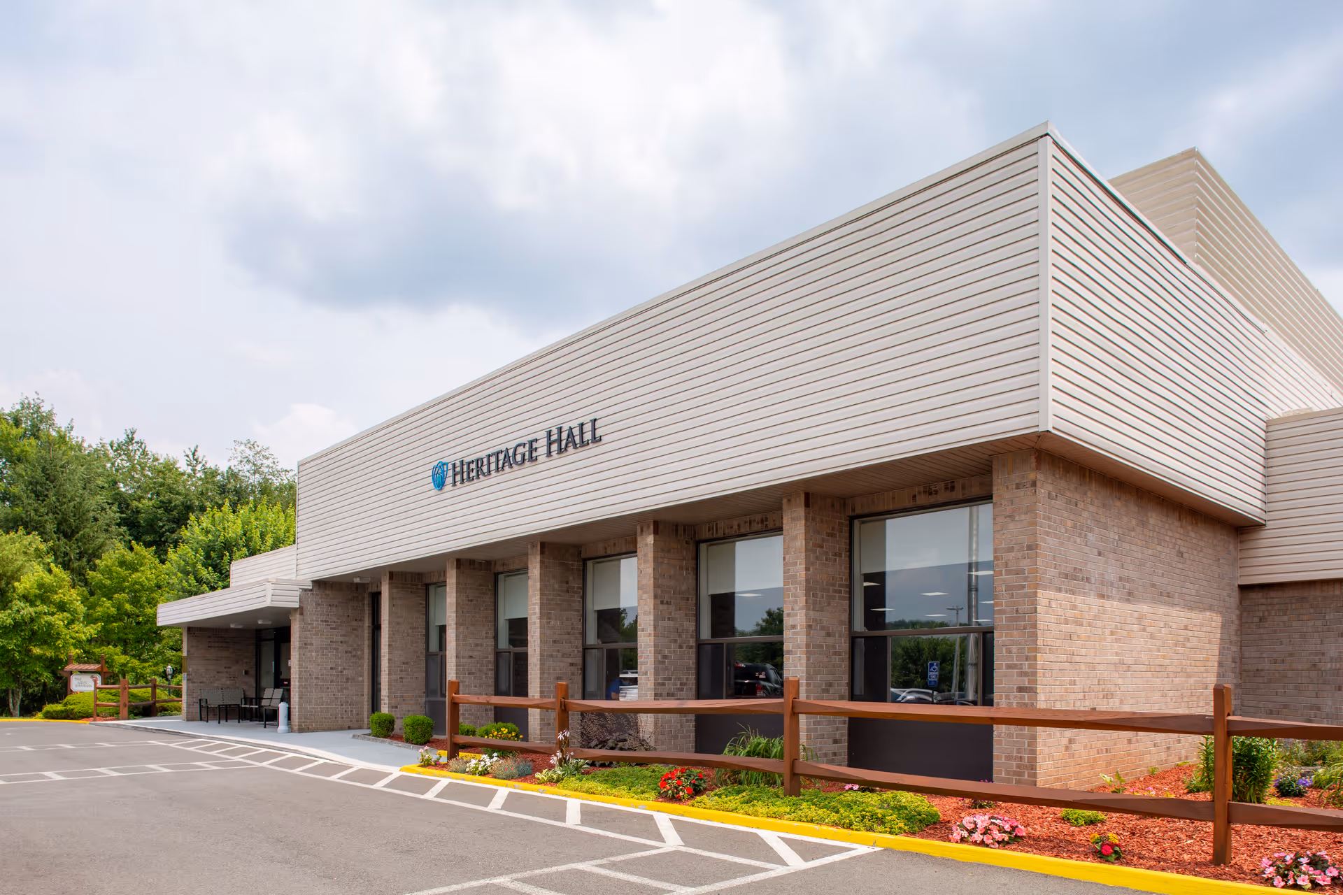 Exterior view of Heritage Hall Tazewell building with brick walls, large windows, a wooden fence, landscaped flower beds, and a parking area under a cloudy sky.