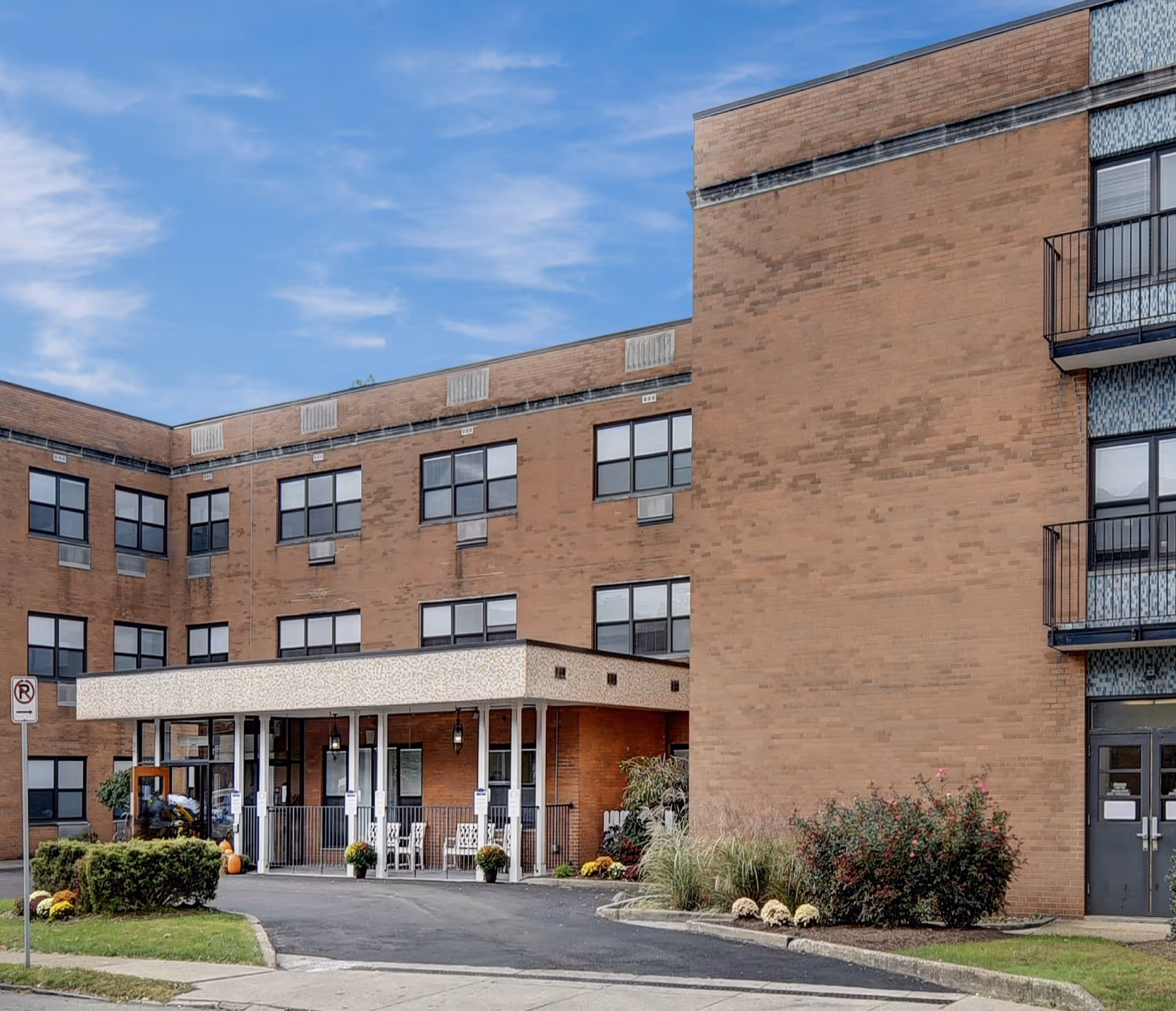 Brick multi-story nursing facility exterior showing a covered entrance, windows, balconies, and a landscaped driveway.