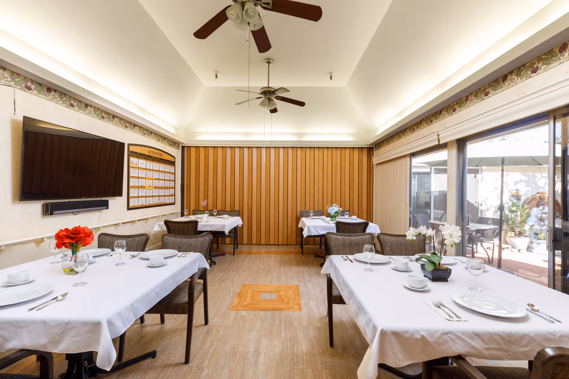 A dining room with several tables covered in white tablecloths, each set with plates, bowls, glasses, and silverware. There are decorative flowers on the tables. The room has a high ceiling with two ceiling fans, a large flat-screen TV mounted on the wall, and a bulletin board labeled 'Activities'. Large sliding glass doors on the right side open to an outdoor patio area with seating and an umbrella.