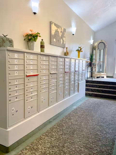 A hallway in a senior living facility with a row of beige mailboxes mounted on the wall. Above the mailboxes are decorative items including a vase with flowers, a bottle, and a world map artwork. The hallway is carpeted with patterned flooring and has wall-mounted lights. In the background, there is a large grandfather clock and a plant on a stand near a set of carpeted stairs.