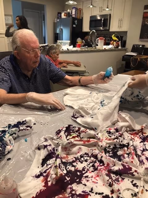Two elderly individuals sitting at a table in a kitchen area, engaging in a tie-dye activity with white shirts and various colors of dye. One person is holding a bottle of blue dye while the other is assisting with the fabric. The kitchen in the background has white cabinets, a refrigerator, and a microwave.