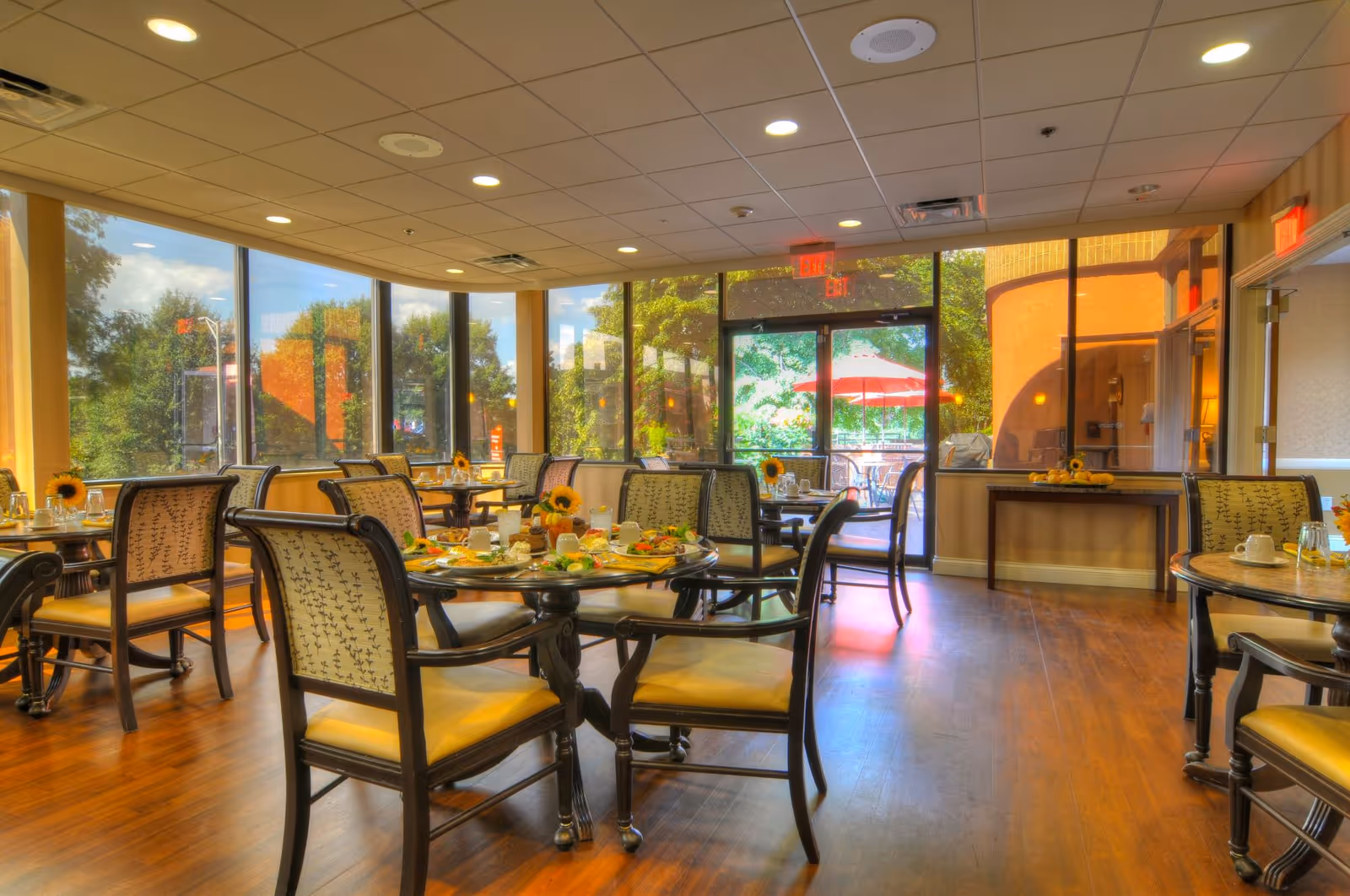 A bright dining room with several round tables set for a meal, each table surrounded by cushioned chairs. Large windows let in natural light and offer a view of greenery and an outdoor patio with red umbrellas. The room has wooden flooring and a ceiling with recessed lighting.