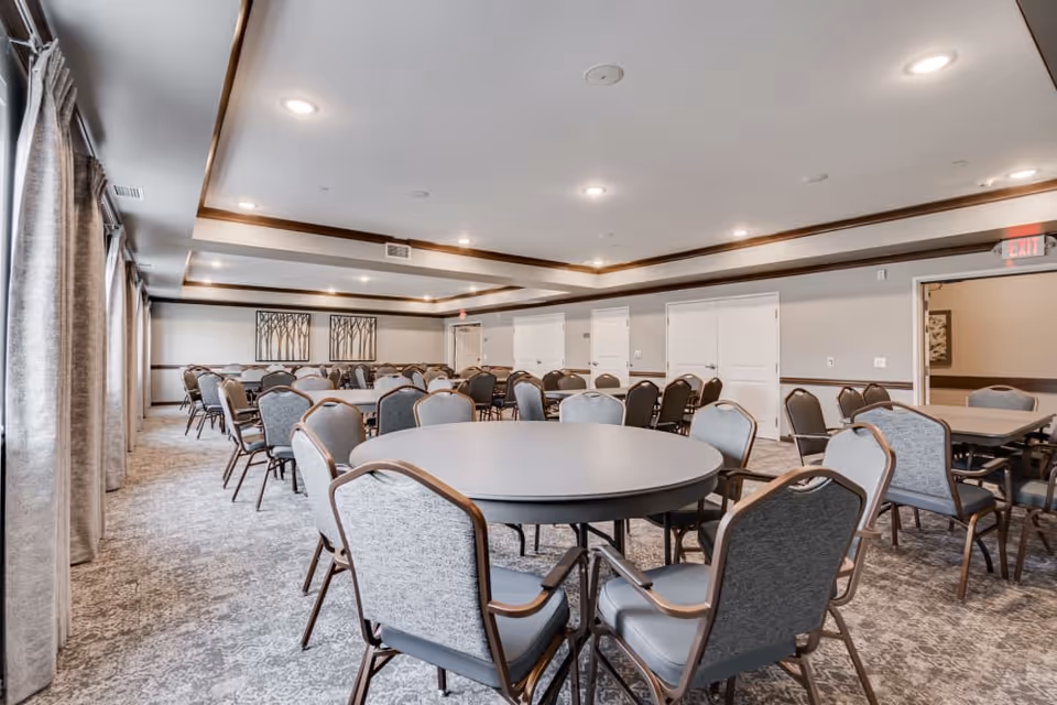 A spacious dining room with multiple round and rectangular tables surrounded by cushioned chairs. The room has a neutral color scheme with gray carpet, light walls, and ceiling lights. Large windows with curtains line one side of the room, and framed artwork is visible on the far wall.