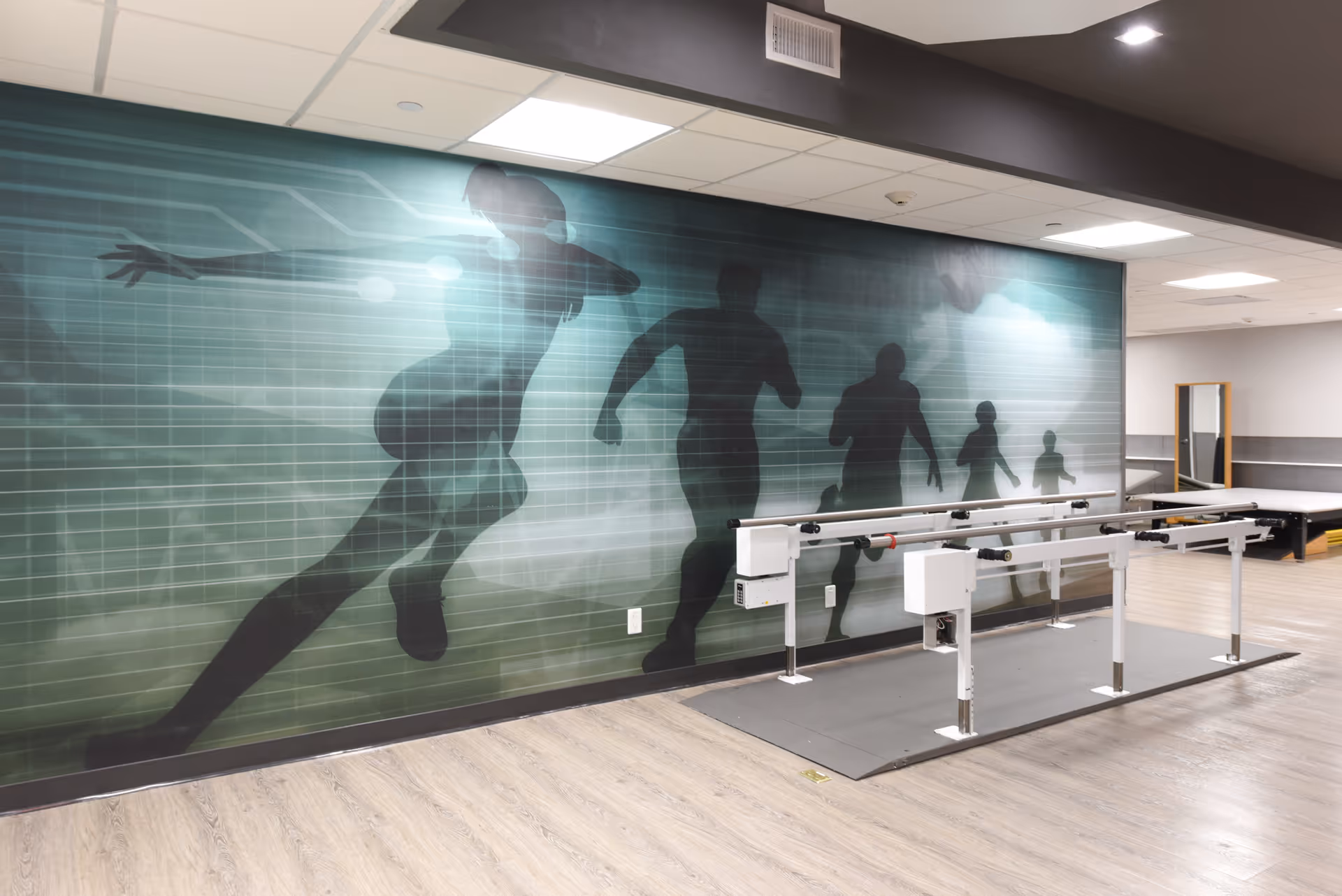 Interior view of a rehabilitation center room with parallel bars for physical therapy. The wall behind the bars features a large mural of silhouettes of people running, depicted in shades of green and black. The room has a light wood floor, a mirror on the far wall, and bright overhead lighting.