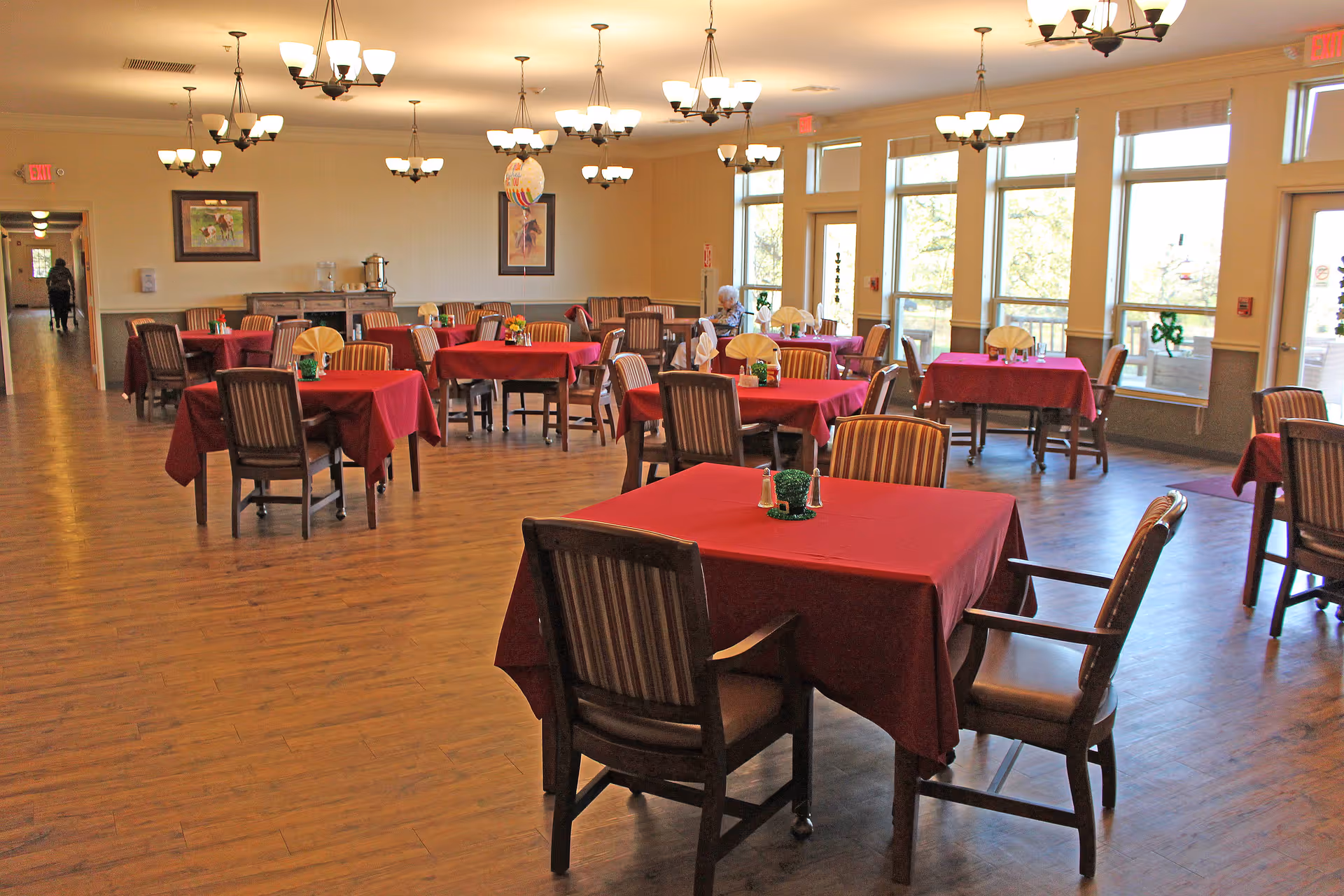Spacious dining room with multiple tables covered in red tablecloths and wooden chairs beneath chandeliers and large windows.