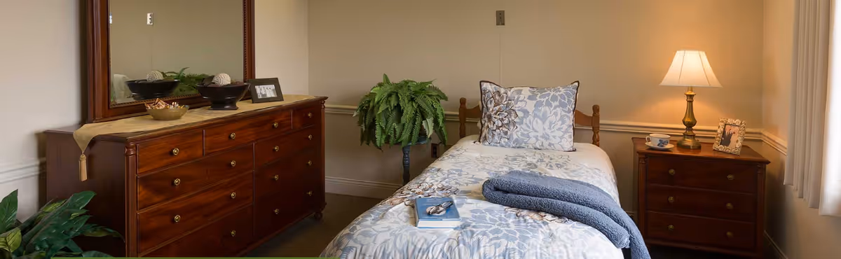 A cozy bedroom with a single bed covered in floral bedding and a folded blue blanket. There is a wooden dresser with a large mirror on the left side, decorated with a bowl, a small plant, and a framed photo. A green potted plant is placed in the corner. On the right side, there is a wooden nightstand with a lit lamp, a teacup and saucer, and a framed photo. Light filters in through a window with white curtains.