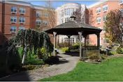 Courtyard of a senior living facility with a wooden gazebo, paved walkway, lawn and a brick multi-story building in the background.