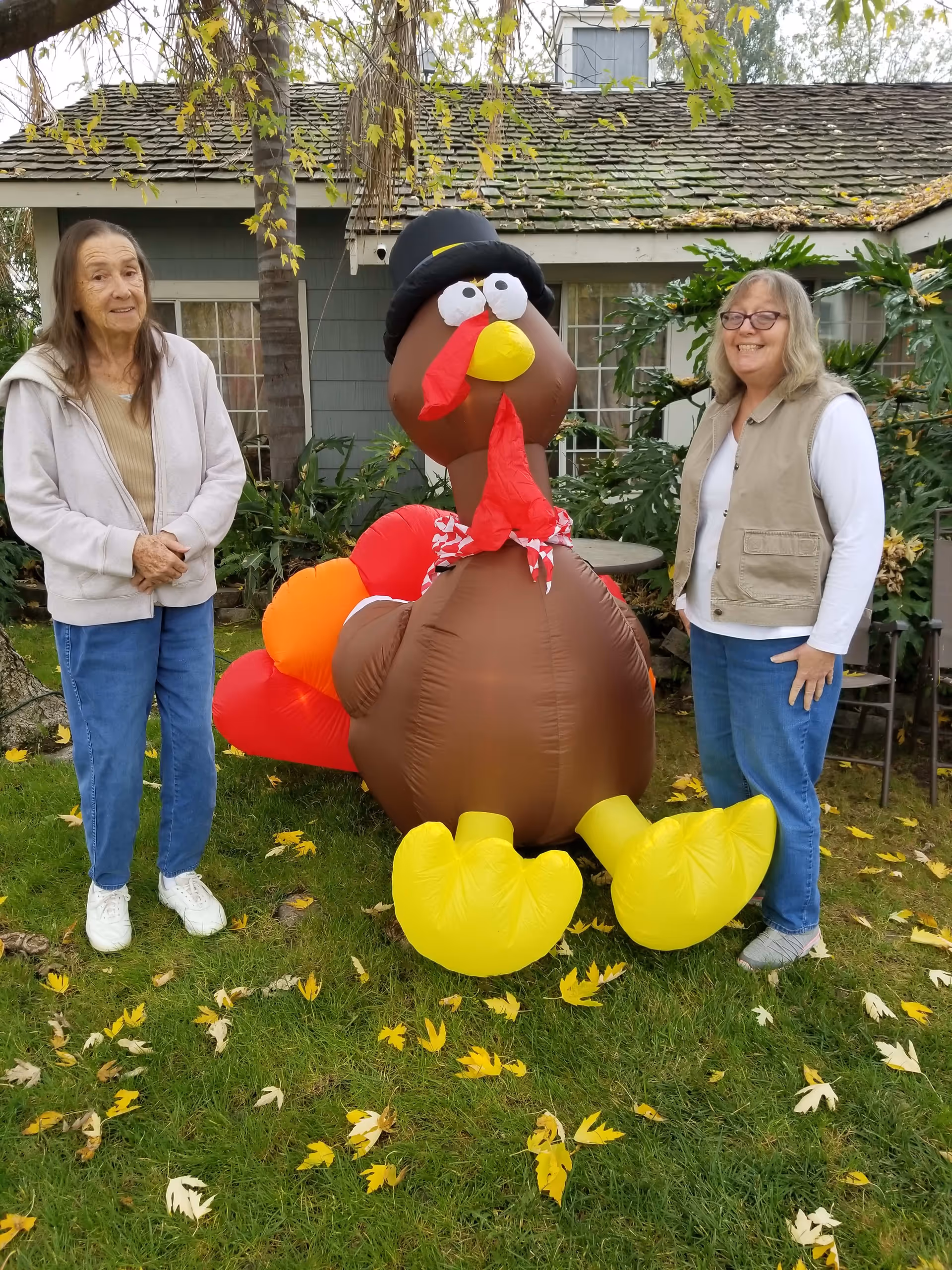 Two elderly women standing outside on grass with fallen leaves, posing next to a large inflatable turkey decoration in front of a house with green siding and windows.