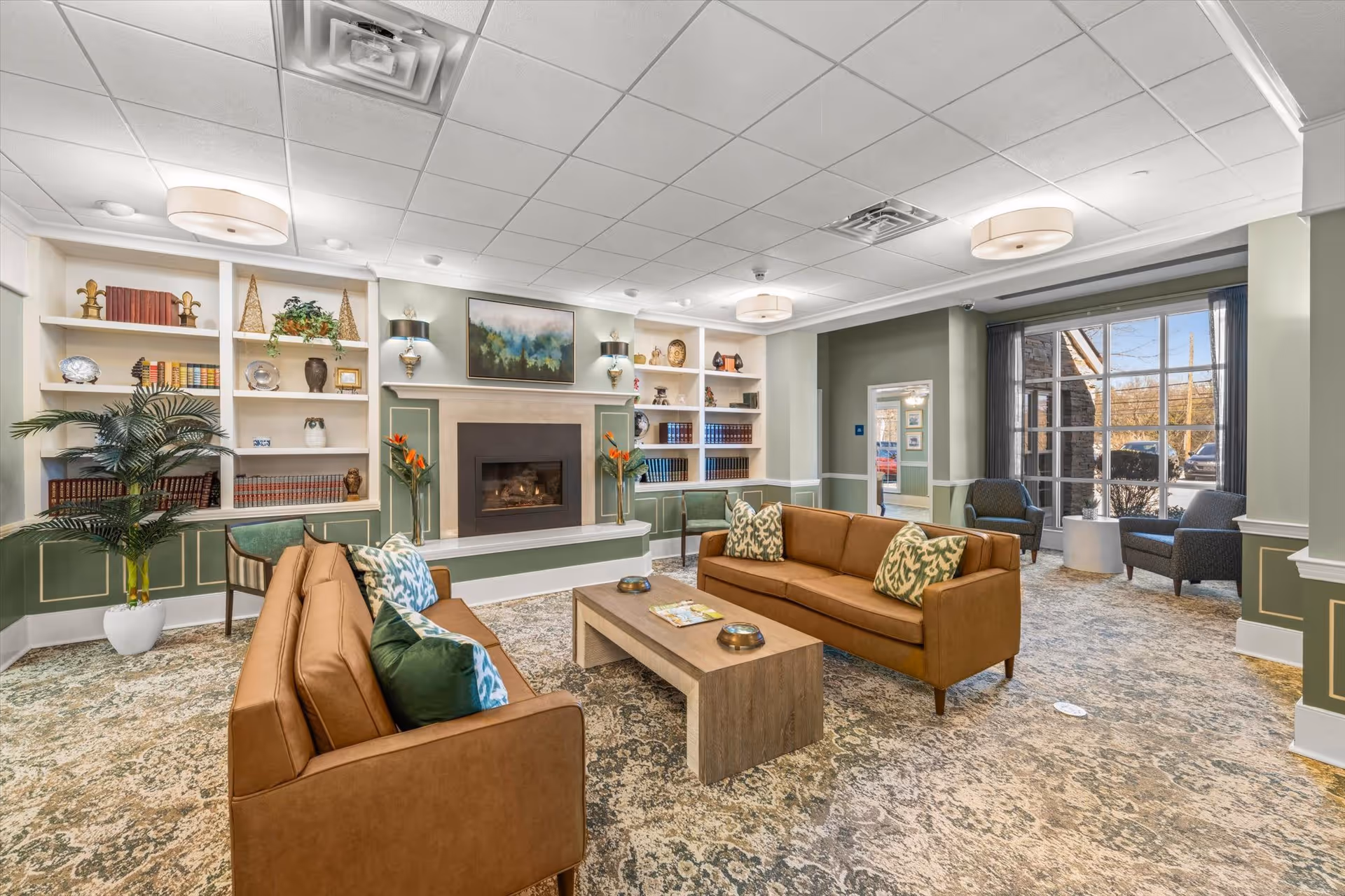 A spacious and well-lit living room with two brown sofas facing each other across a wooden coffee table. The room features a fireplace centered on the far wall, flanked by built-in white shelves filled with books and decorative items. There are two armchairs near a large window letting in natural light, and the walls are painted in a soft green with white trim. The carpet has a patterned design, and ceiling lights provide additional illumination.
