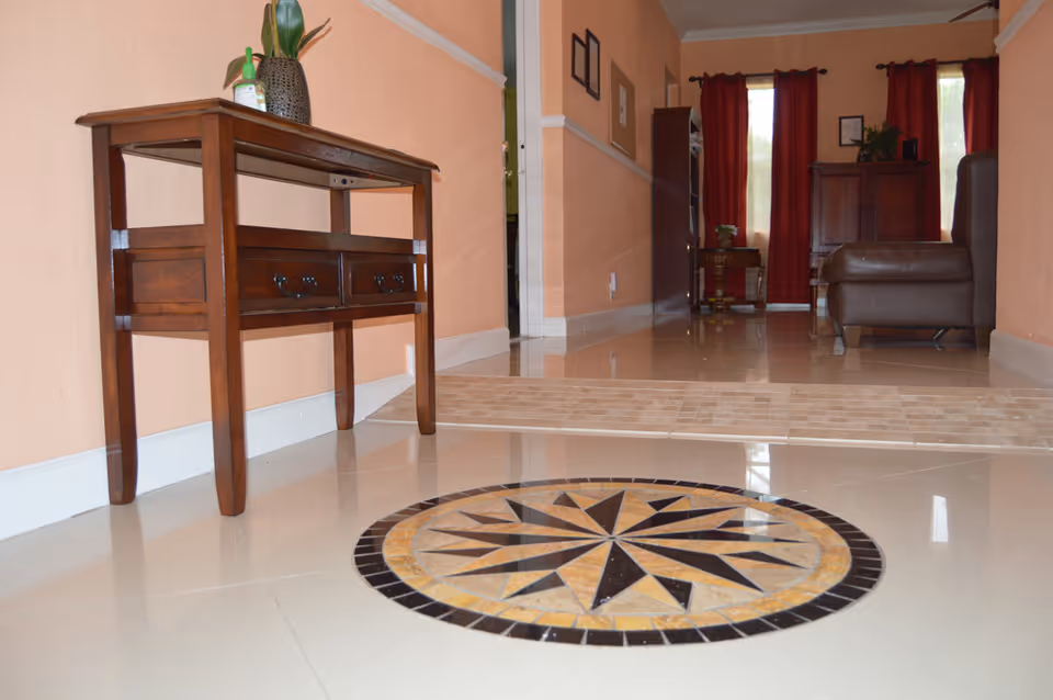 Interior hallway of a facility with peach-colored walls and shiny tiled floor featuring a circular decorative tile pattern. A wooden console table with two drawers and a plant on top is positioned against the left wall. In the background, there is a living area with red curtains, a brown leather chair, and wooden furniture.