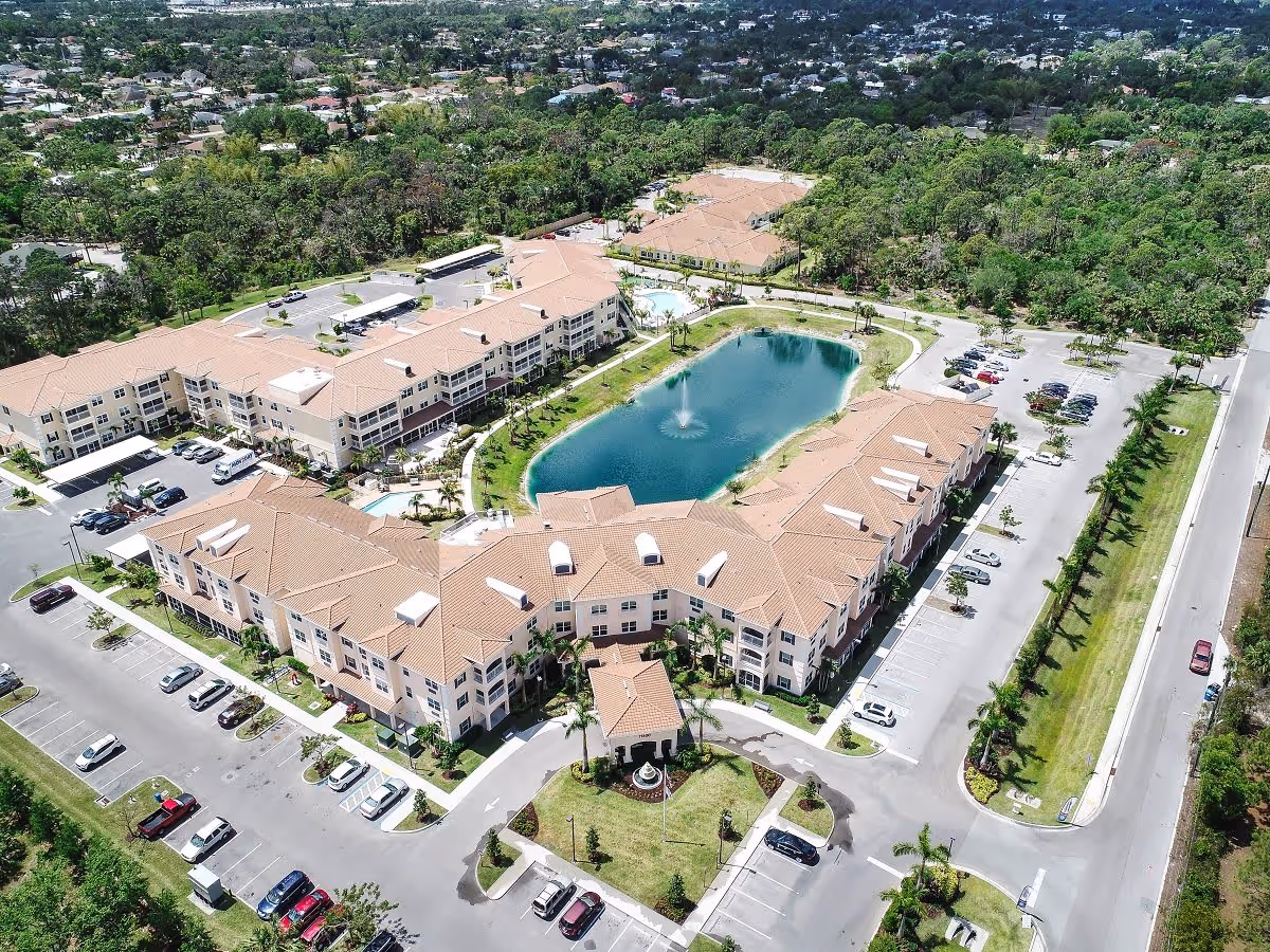 Aerial view of the American House Bonita Springs senior living facility showing multiple beige buildings with terracotta roofs arranged around a central pond with a fountain. The facility is surrounded by parking lots, green lawns, and trees, with a road running alongside the property.