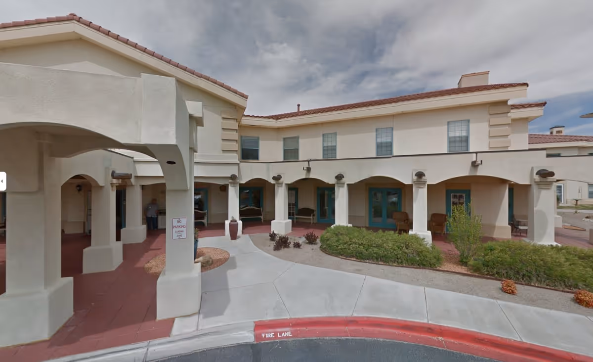 Exterior view of a two-story senior living facility building with beige stucco walls and a red tile roof. The entrance area features covered walkways with arches and columns, outdoor seating, and landscaped bushes. A red curb marked 'FIRE LANE' and a 'NO PARKING LOADING ZONE' sign are visible near the entrance.