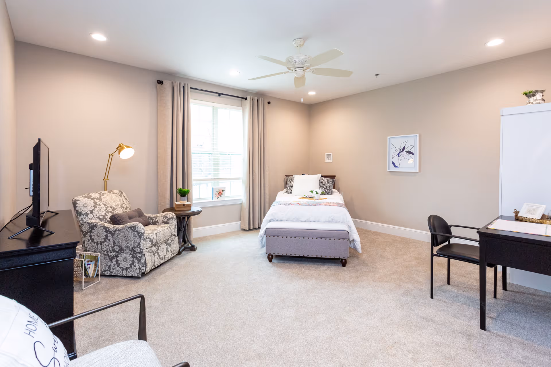 Bright, spacious assisted-living bedroom with a bed against the back wall, an upholstered armchair by a window, a TV on a dresser, and a small desk.