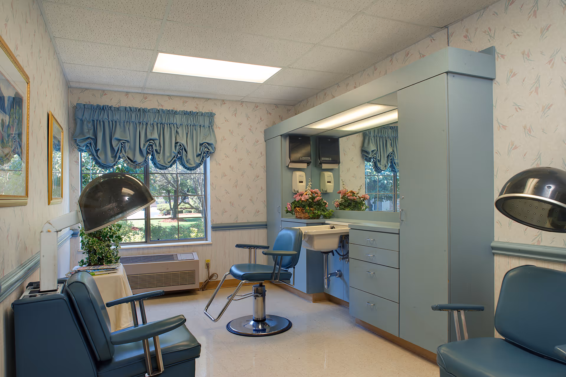 Small salon room with blue styling chairs, hooded hair dryers, a mirrored vanity with sink and floral decor, and a window with a blue valance.