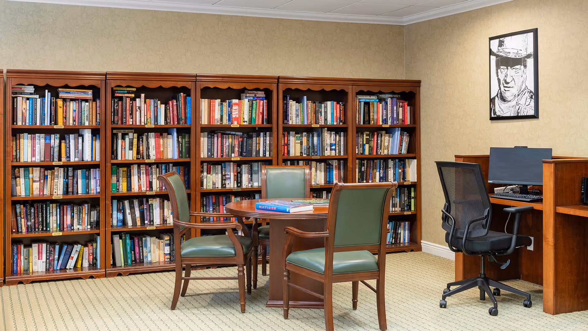 A cozy library room with wooden bookshelves filled with books along the back wall. In front of the shelves is a round wooden table surrounded by four green cushioned wooden chairs. To the right, there is a wooden desk with a computer and a black mesh office chair. A black and white framed portrait hangs on the beige wall above the desk.