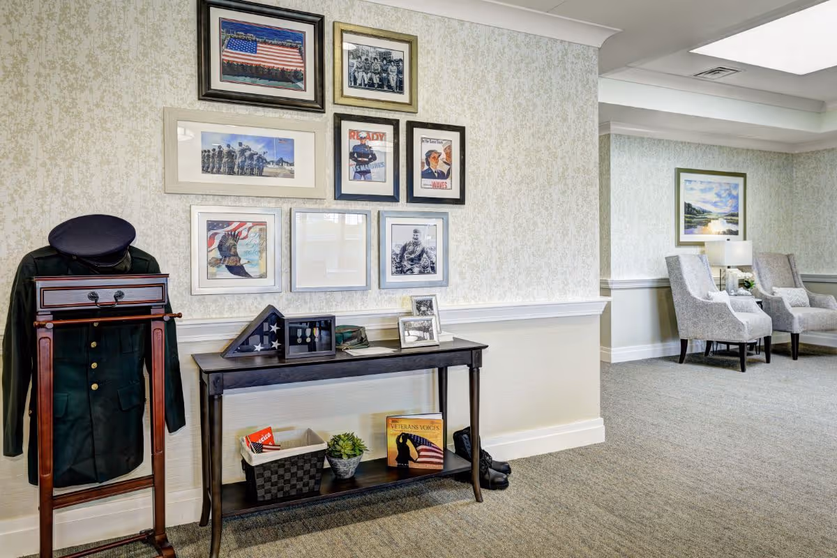 A hallway corner in a senior living facility featuring a military uniform on a wooden valet stand, a console table with framed photos, a folded American flag in a display case, a basket, a small plant, and a book titled 'Veterans Voices'. The wall above the table displays framed patriotic and military-themed pictures. In the background, there are two upholstered armchairs with a side table and lamp between them.