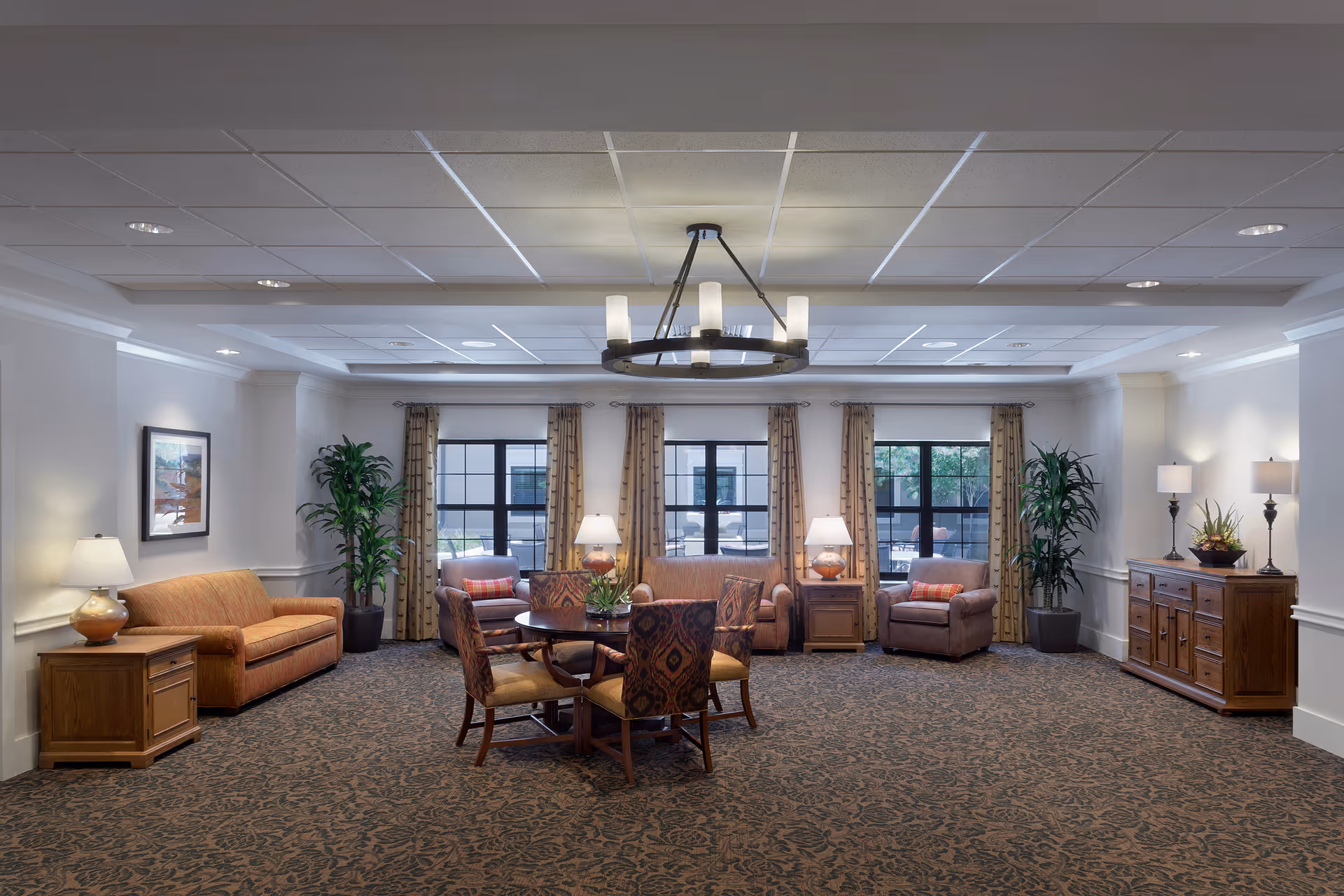 A spacious senior living common area with patterned carpet, a round wooden table with four upholstered chairs in the center, two armchairs and a sofa along the back wall, large windows with curtains, potted plants, and multiple lamps providing warm lighting.