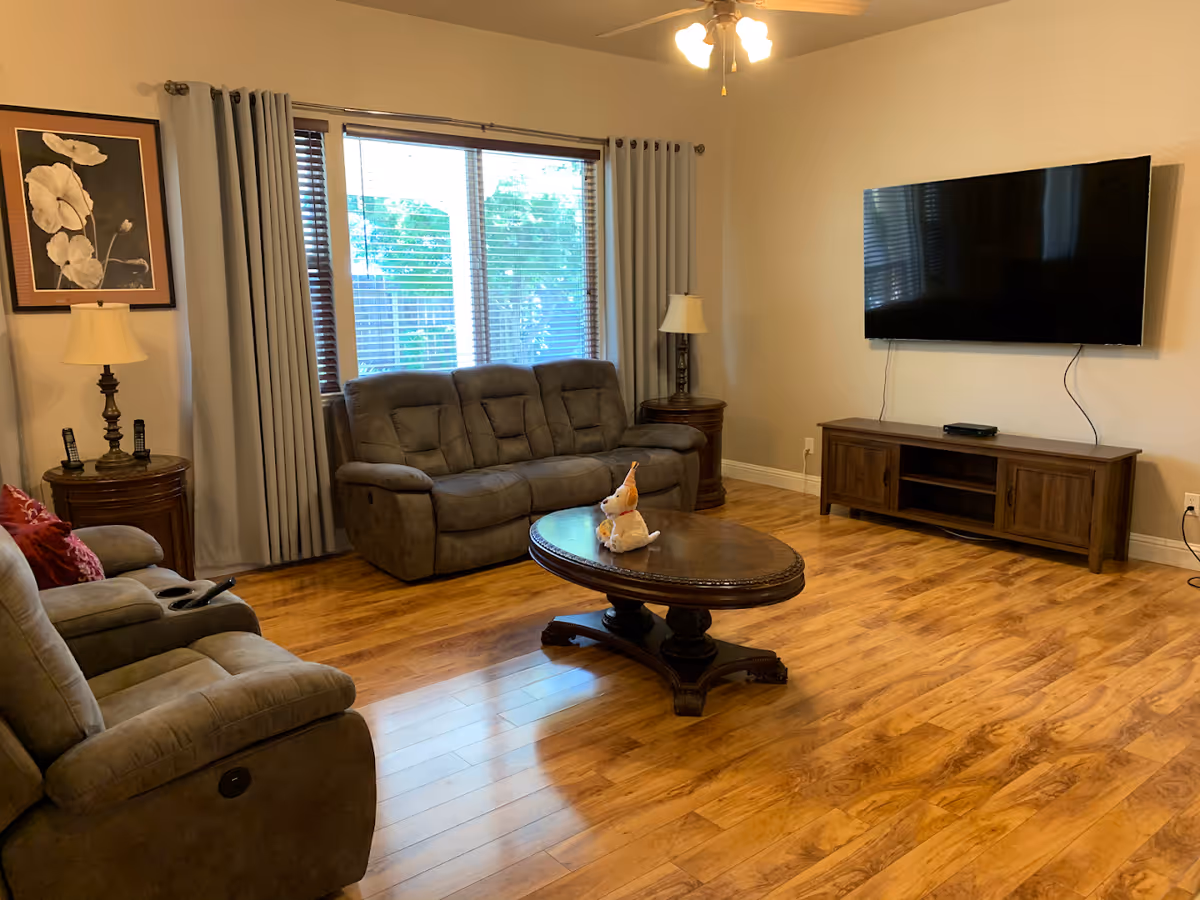A cozy living room with wooden flooring, a large window with gray curtains, a gray reclining sofa and armchair, two side tables with lamps, a wooden coffee table with a small stuffed animal on it, and a flat-screen TV mounted on the wall above a wooden TV stand.