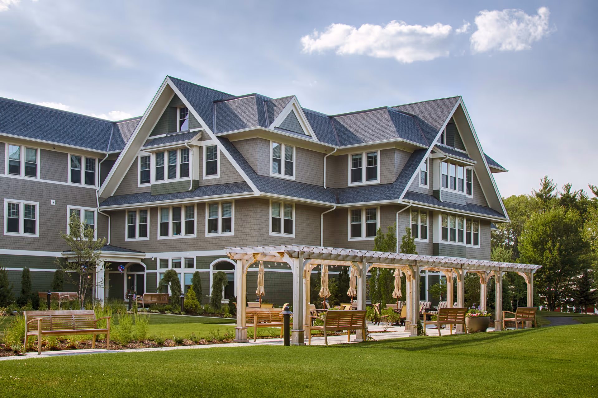 Exterior view of a large, multi-story residential building with gray siding and white trim. In front of the building is a well-maintained lawn with wooden benches and a pergola structure with hanging umbrellas. Trees and shrubs surround the area under a partly cloudy sky.