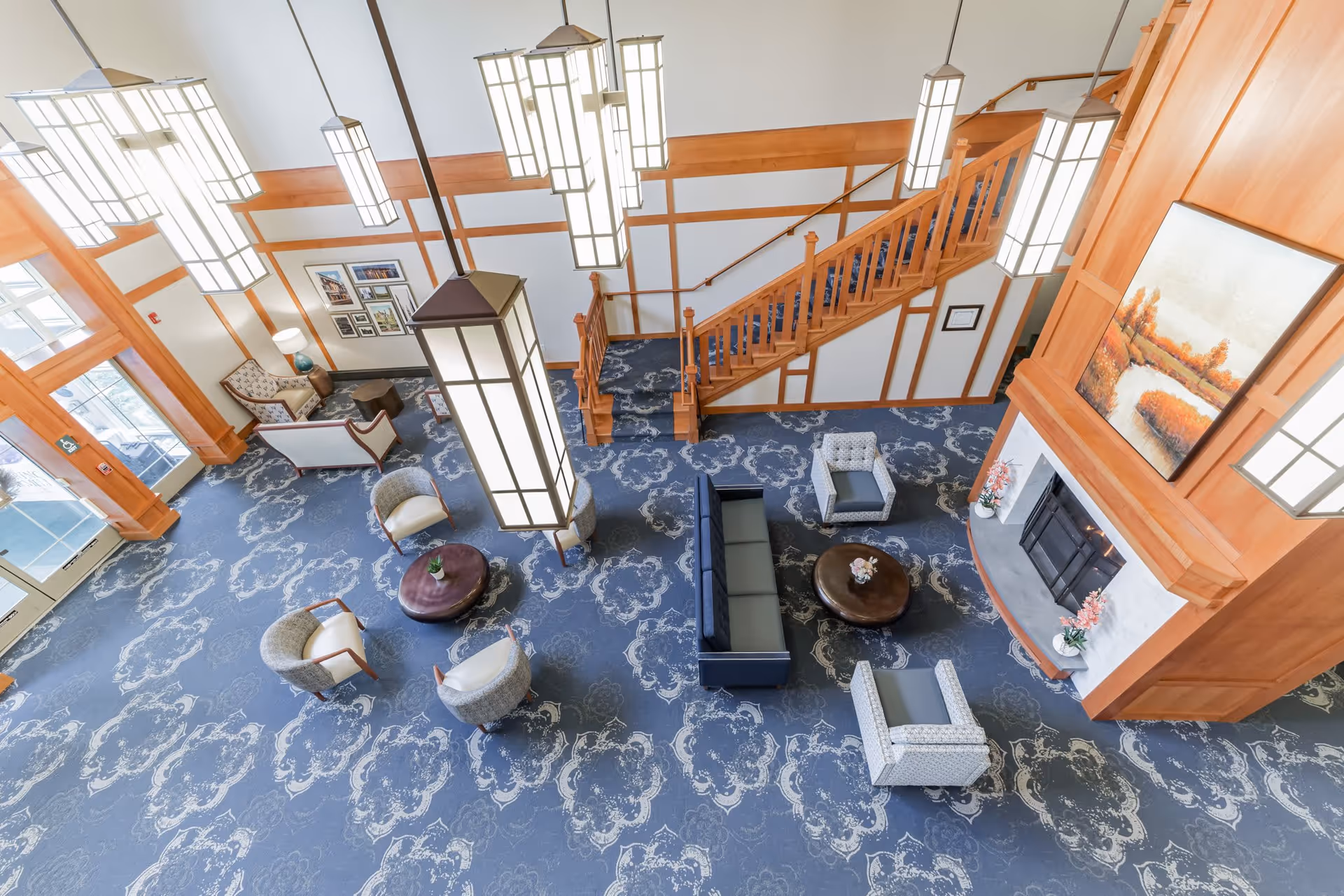 A spacious and well-lit living room area with a blue patterned carpet, multiple seating arrangements including armchairs and sofas, a wooden staircase, large hanging pendant lights, and a fireplace with a landscape painting above it.