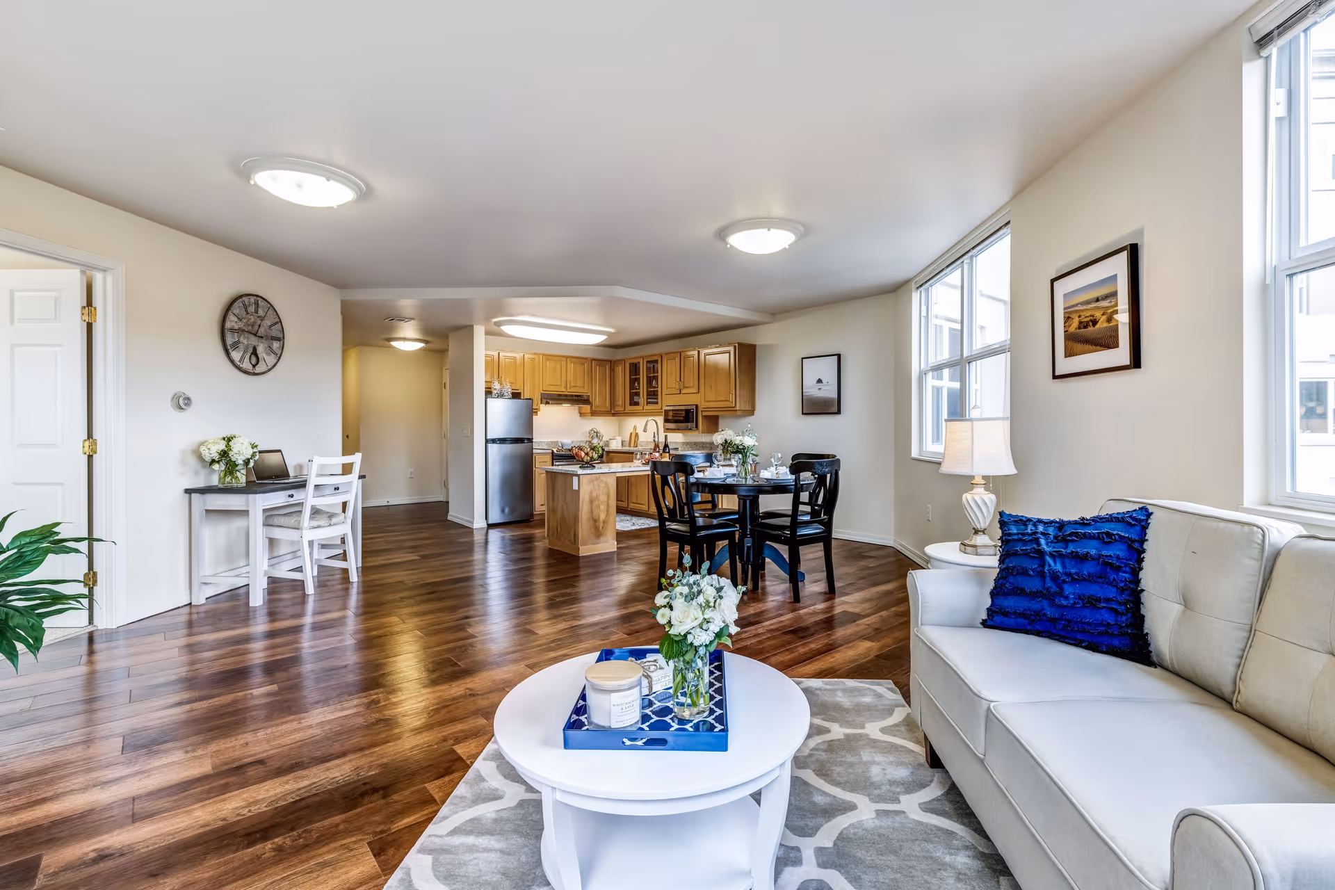Bright and spacious open-concept living area in a senior living facility featuring a light gray sofa with a blue pillow, a white round coffee table with a flower arrangement and candle, a dining table with four black chairs, and a kitchen with wooden cabinets and stainless steel appliances. The room has large windows letting in natural light, wooden flooring, and neutral-colored walls with framed artwork.
