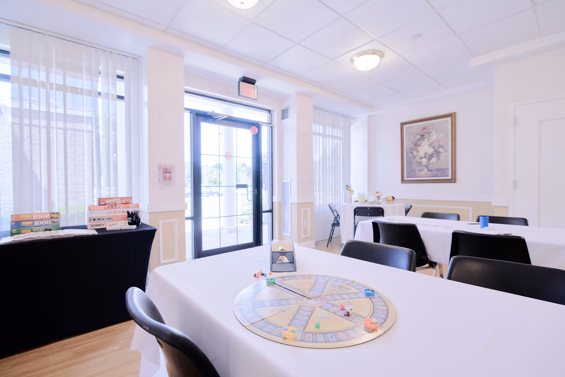 Bright community room with tables covered in white cloths and a board game on the foreground table near a glass exit door.