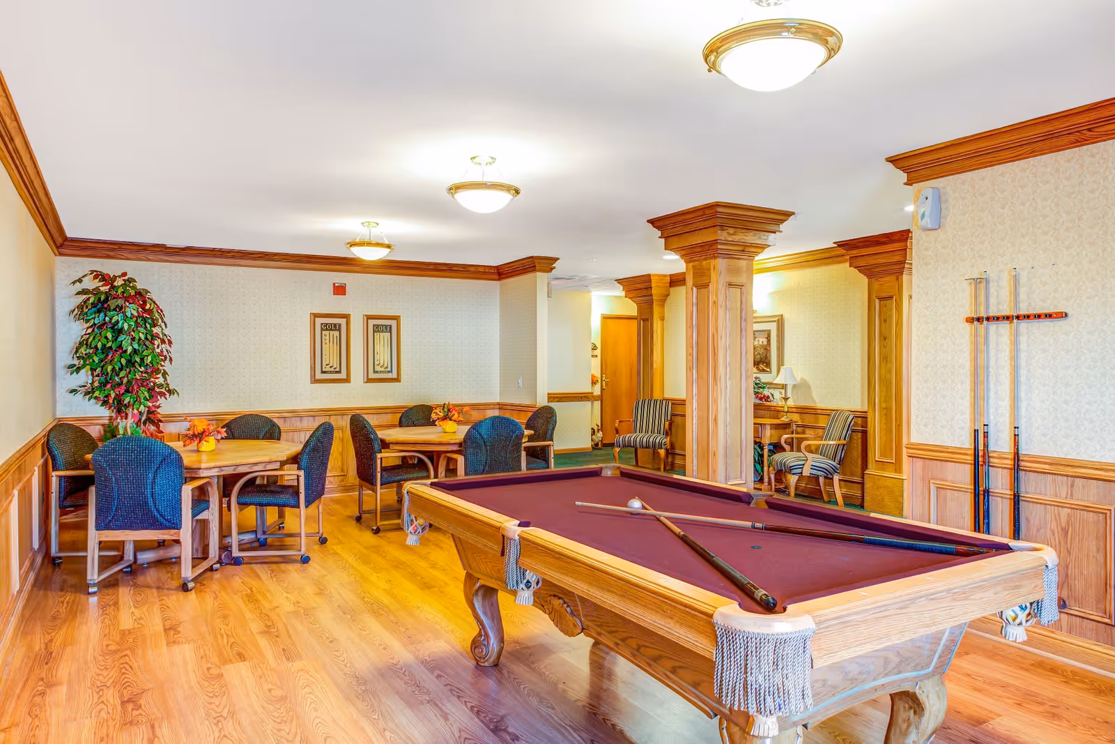 A recreational room with a pool table in the foreground, two pool cues resting on it, and a cue rack on the wall. The room has wooden flooring, wood-paneled walls, and several round tables with chairs in the background. There are also two framed pictures on the far wall, a potted plant, and two striped armchairs near a wooden column.