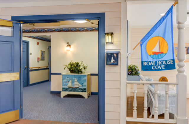 View of an interior hallway with blue doors and trim, a small chest painted with a sailboat, a plant on top, and a wall light. To the right, a porch area with white wicker furniture and a blue flag with a sailboat and the words 'Boat House Cove'.