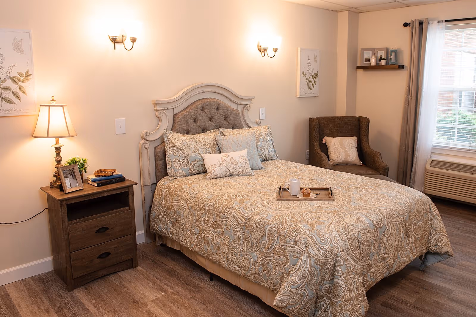 A cozy bedroom with a large bed featuring a patterned beige and blue comforter and multiple pillows. There is a wooden nightstand with a lamp, books, a small plant, and a framed photo. A cushioned armchair with a pillow is placed near a window with white curtains and an air conditioning unit below it. The walls are decorated with framed botanical prints and a small shelf with decorative items.