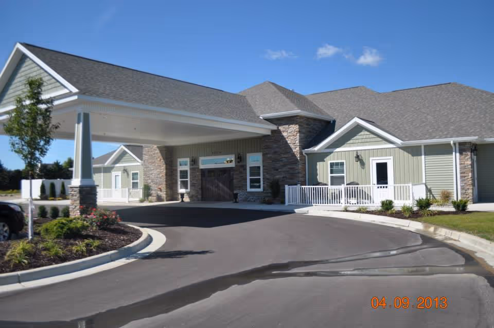 Entrance of a single-story assisted living facility with a covered porte-cochere, stone accents, and landscaped driveway under a blue sky.