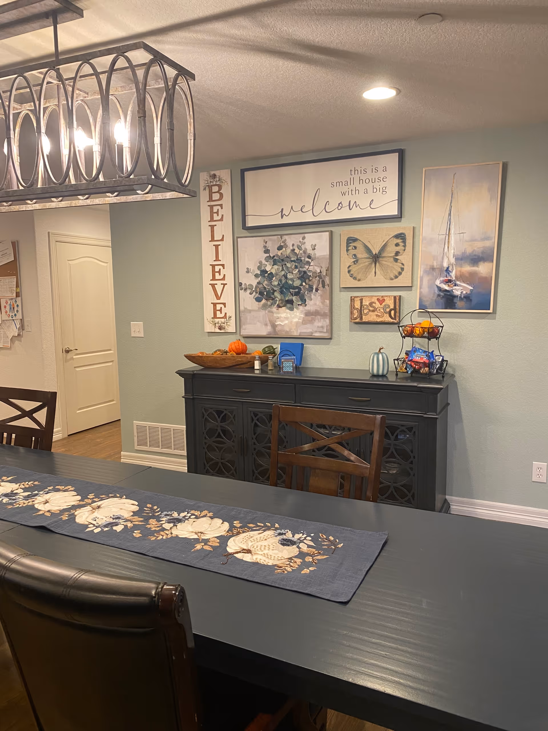 A dining room with a dark wooden table and chairs. The table has a blue table runner with a white floral pattern. On the wall behind the table is a black sideboard with decorative items including a bowl with small pumpkins and a tiered basket with snacks. Above the sideboard are several framed artworks and signs, including one that says 'this is a small house with a big welcome', a vertical sign that says 'BELIEVE', a painting of a butterfly, a floral painting, and a painting of a sailboat. A modern chandelier with exposed bulbs hangs above the table.