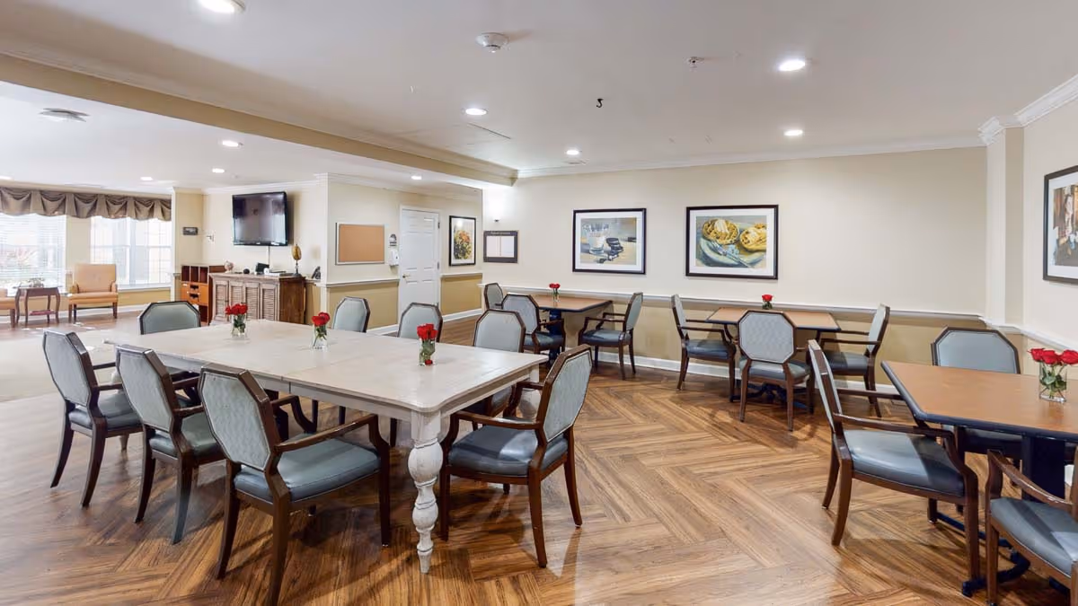 A well-lit dining room in a senior living facility with multiple tables and chairs arranged neatly. Each table has a small vase with red flowers. The room features wood-patterned flooring, beige walls adorned with framed artwork, and a television mounted on one wall. Large windows allow natural light to enter, and there is a seating area with armchairs near the windows.