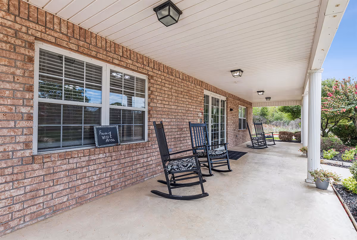 Covered porch area with brick walls and white ceiling, featuring three black rocking chairs with patterned cushions. A small chalkboard sign on the window reads 'Family Visit Area'. The porch overlooks a garden with green shrubs and flowering plants.