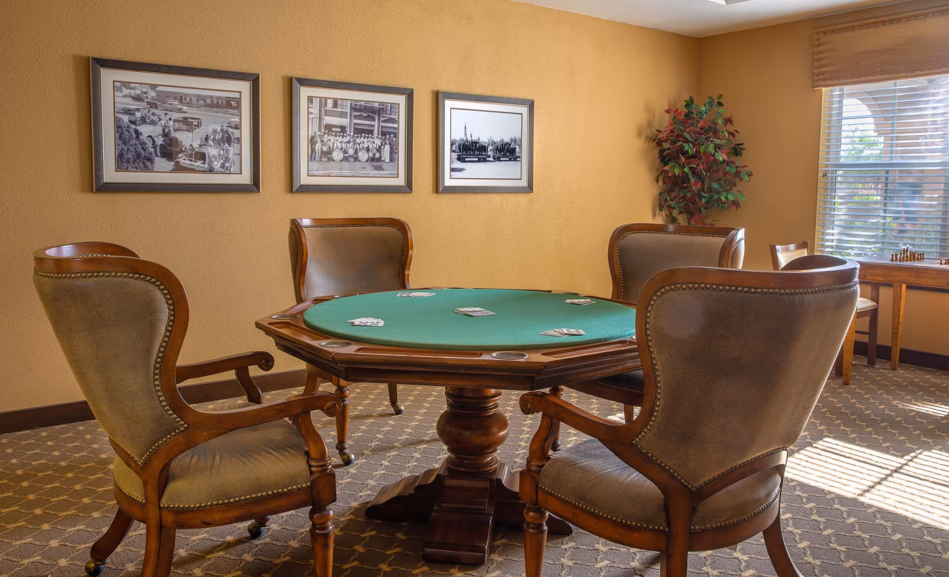 A cozy game room with a round wooden poker table covered with a green felt surface and playing cards placed on it. Four upholstered wooden chairs surround the table. The room has beige walls adorned with three framed black and white photographs. A window with blinds allows natural light to brighten the space, and a small table with a chessboard is visible near the window. A potted plant adds a touch of greenery to the corner.