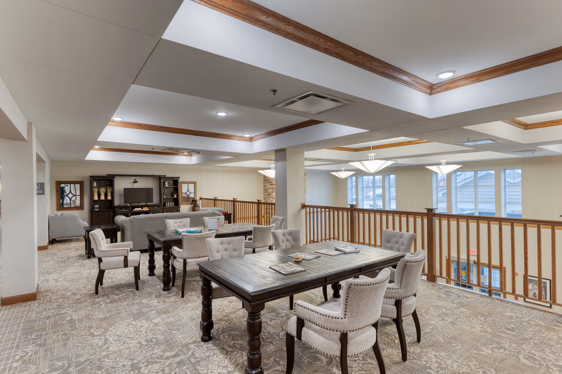 A spacious senior living common area with a large wooden table surrounded by six upholstered chairs in the foreground. Behind it, there is a seating area with sofas and armchairs facing a TV on a dark wooden entertainment center. The room has beige walls, patterned carpet, wooden ceiling trim, and large windows letting in natural light. The area is on an upper floor with a wooden railing overlooking the lower level.