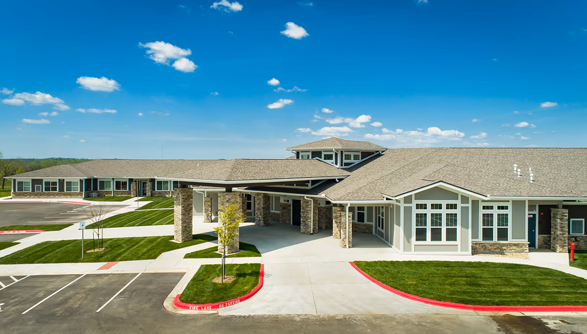 Exterior view of a single-story assisted living and memory care facility building with a covered entrance, stone pillars, large windows, and neatly maintained green lawns under a bright blue sky with scattered clouds.