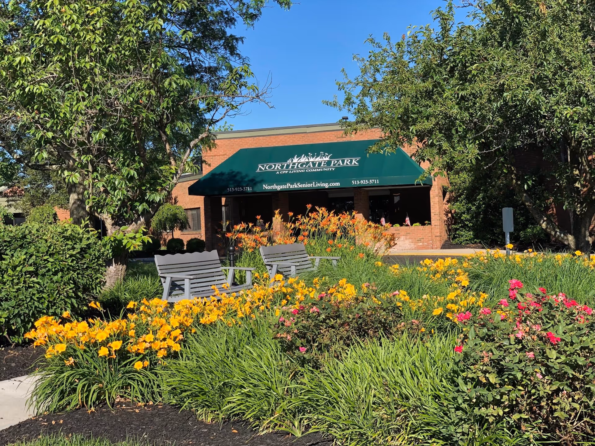 Outdoor garden area with blooming yellow and pink flowers, green shrubs, and two gray benches in front of a brick building with a green awning that reads 'Northgate Park A CPE Living Community' along with a website and phone number.