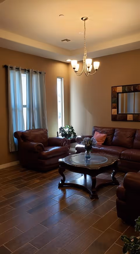A cozy living room with brown leather armchairs and a matching sofa arranged around a wooden coffee table with a glass top. A vase with flowers is on the table. The room has brown tiled flooring, beige walls, a window with light blue curtains, a narrow vertical window, a decorative mirror on the wall, and a ceiling light fixture with five bulbs.
