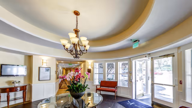 Bright and welcoming facility entrance area with a round glass table holding a large floral arrangement in the center. There is a chandelier hanging from a recessed circular ceiling. To the right, double glass doors lead outside, and a red bench is positioned near the windows. A hallway extends from the entrance area, with wall sconces and framed artwork on the walls.