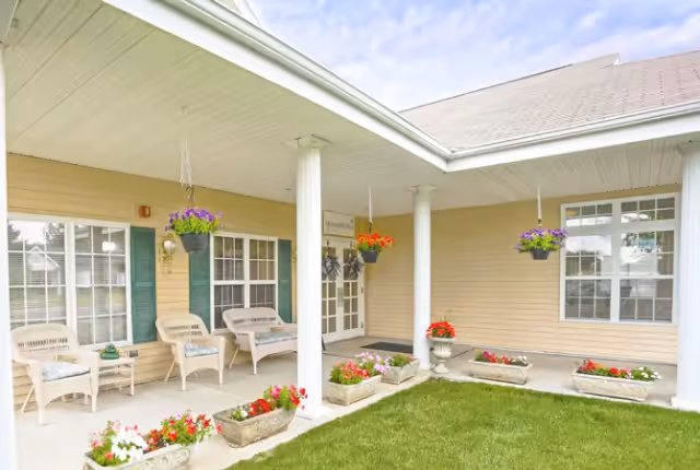 Covered front porch with white columns, wicker chairs, hanging flower baskets and planters along a lawn.