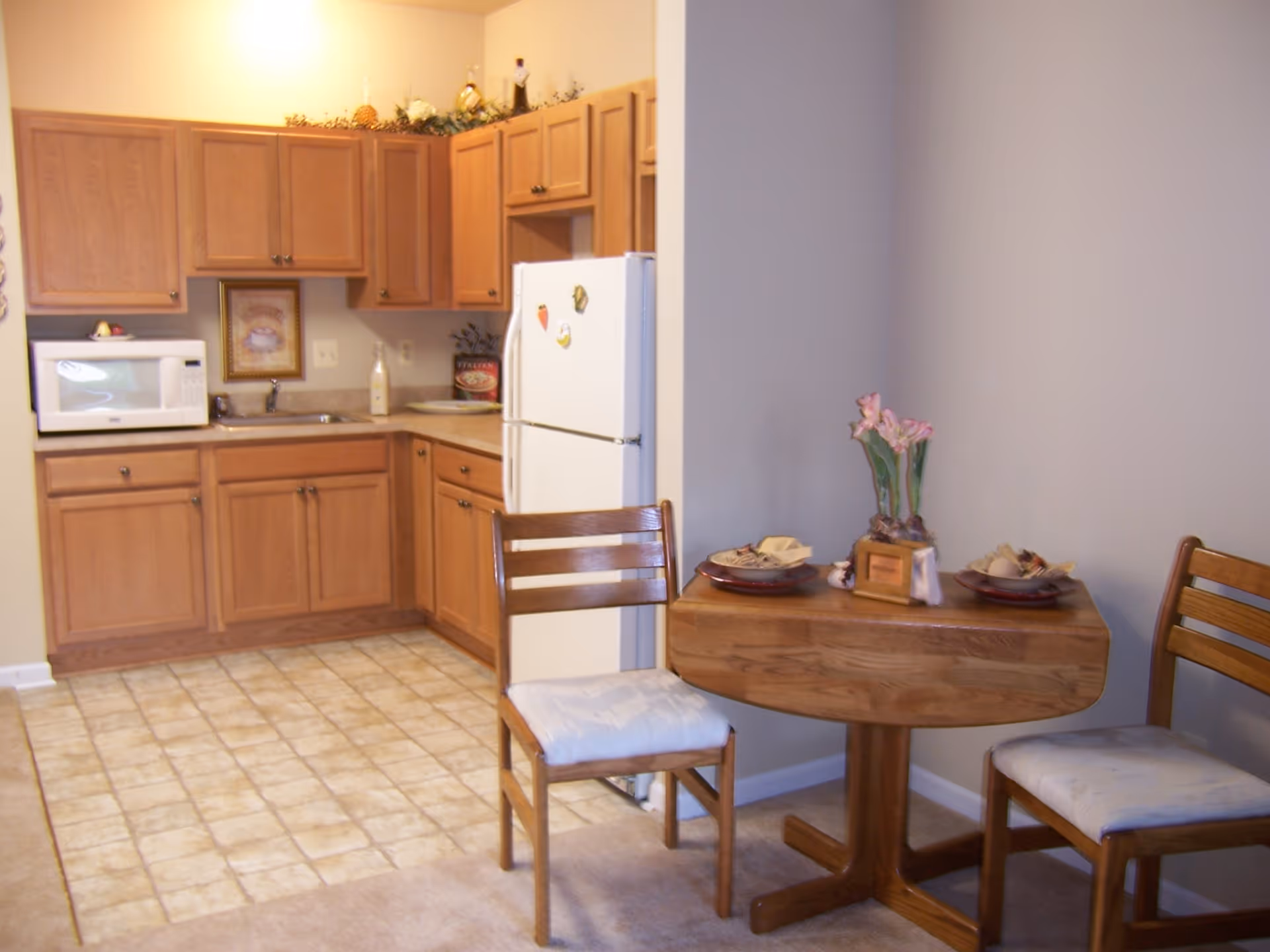 Small kitchen area with wooden cabinets, a white refrigerator, and a microwave on the counter. Adjacent to the kitchen is a small wooden dining table with two chairs, set with plates and napkins, and a small flower arrangement in the center.