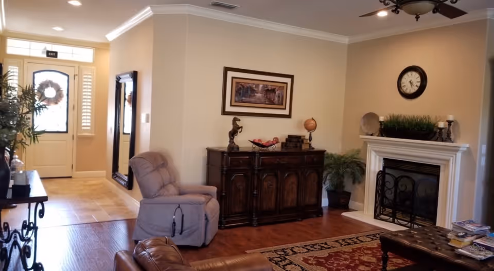 A cozy living room area with a beige recliner chair, a dark wooden cabinet adorned with decorative items including a horse statue and a globe, a fireplace with a clock above it, and a large mirror on the wall near the entrance door. The room has wooden flooring, a patterned rug, and plants adding greenery.