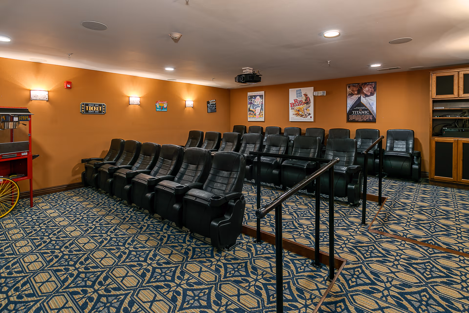A small theater room with two rows of black leather recliner chairs arranged on a carpeted floor with a blue and beige geometric pattern. The walls are painted orange and decorated with movie posters and small wall lights. There is a popcorn machine on the left side and a wooden cabinet with electronic equipment on the right side. A projector is mounted on the ceiling.