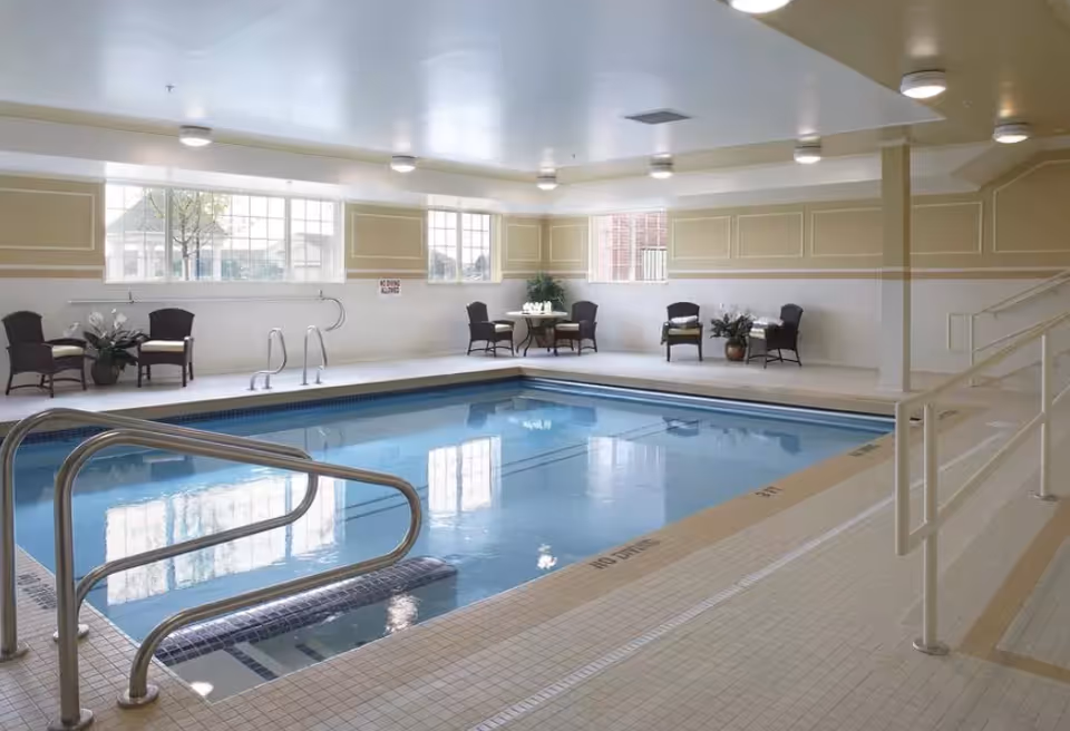 Indoor swimming pool with handrails, seating area, and large windows in a light-filled room.