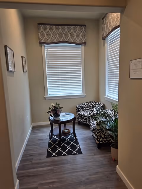 A small sitting area with two patterned armchairs and a round wooden table between them. The table has a flower arrangement and a book on the lower shelf. The floor has a dark patterned rug, and the walls are light-colored with two windows covered by blinds and decorative valances. There are framed pictures on the walls and a potted plant near one of the chairs.