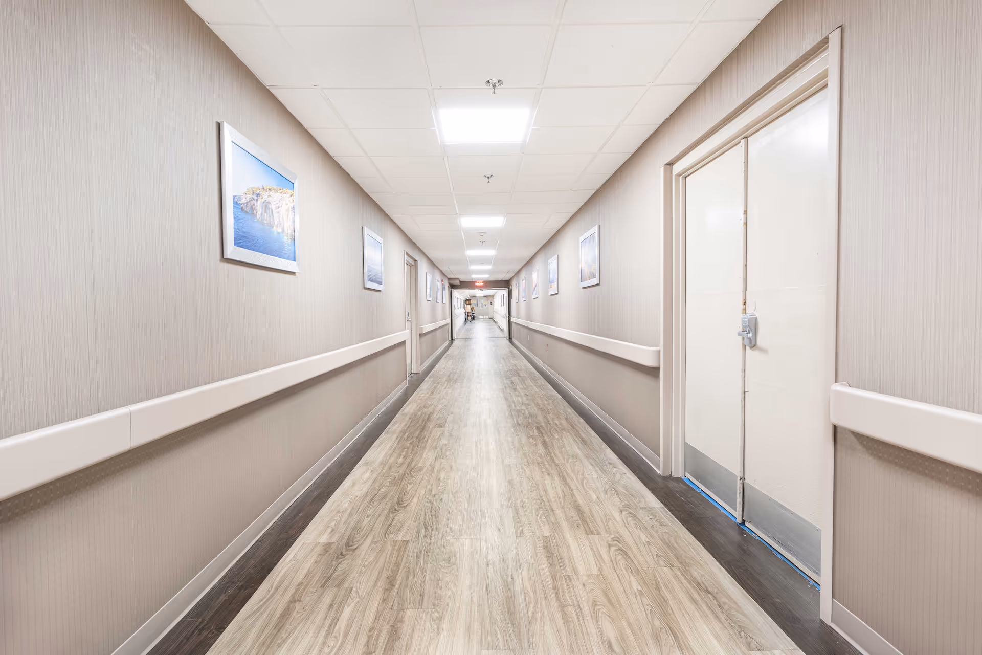 A long, well-lit hallway in a nursing and rehabilitation facility with light wood flooring, beige walls, handrails on both sides, and framed pictures hanging along the walls. Doors line the hallway, and the ceiling has recessed lighting panels.