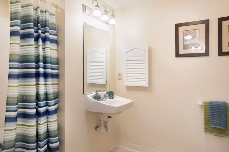 A bathroom with a white wall-mounted sink, a rectangular mirror above it, and a light fixture with three bulbs. To the left, there is a shower with a striped shower curtain in shades of blue, green, and white. On the right wall, there are two framed pictures and a towel rack holding two towels, one green and one blue. A white wall cabinet with a louvered door is mounted above the sink.
