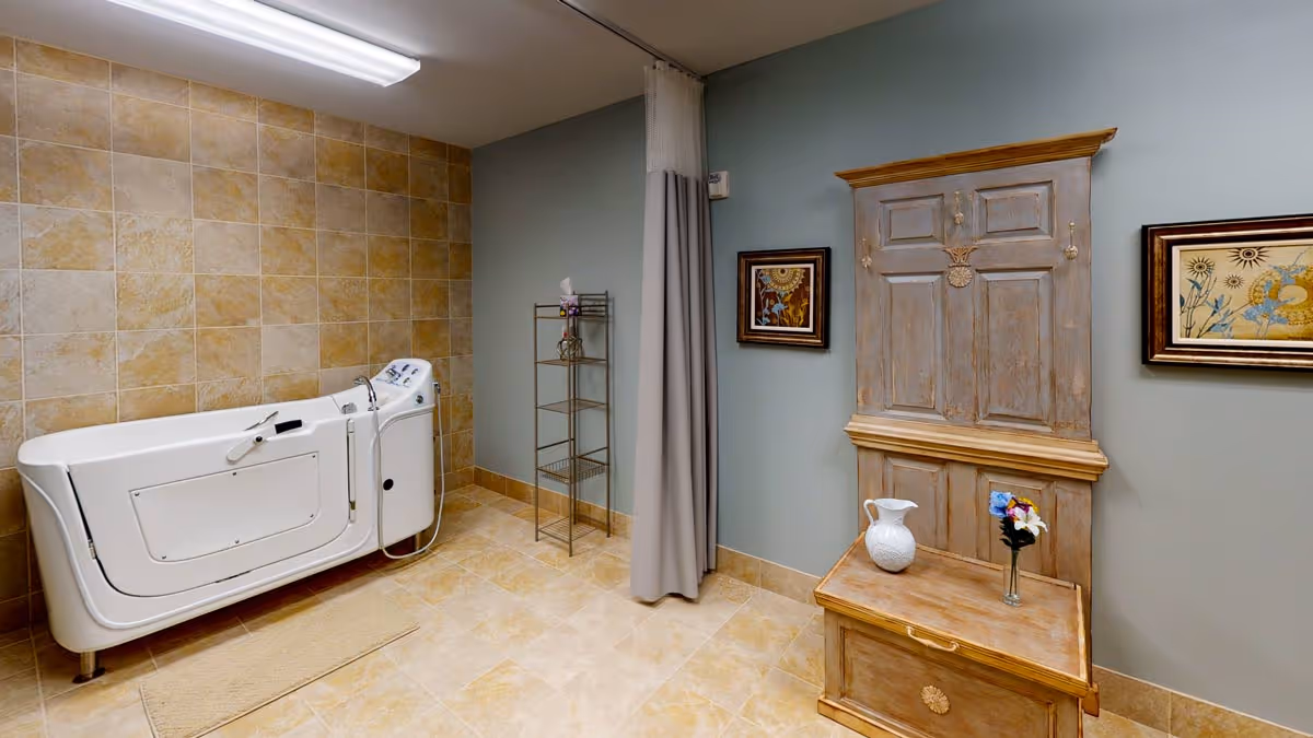 A spacious bathroom with beige tiled walls and floor featuring a white walk-in bathtub on the left side. On the right side, there is a wooden cabinet with hooks and a small bench, decorated with a white pitcher and a glass vase with flowers. Two framed pictures hang on the wall above the cabinet. A metal shelving unit stands near a gray curtain in the corner.