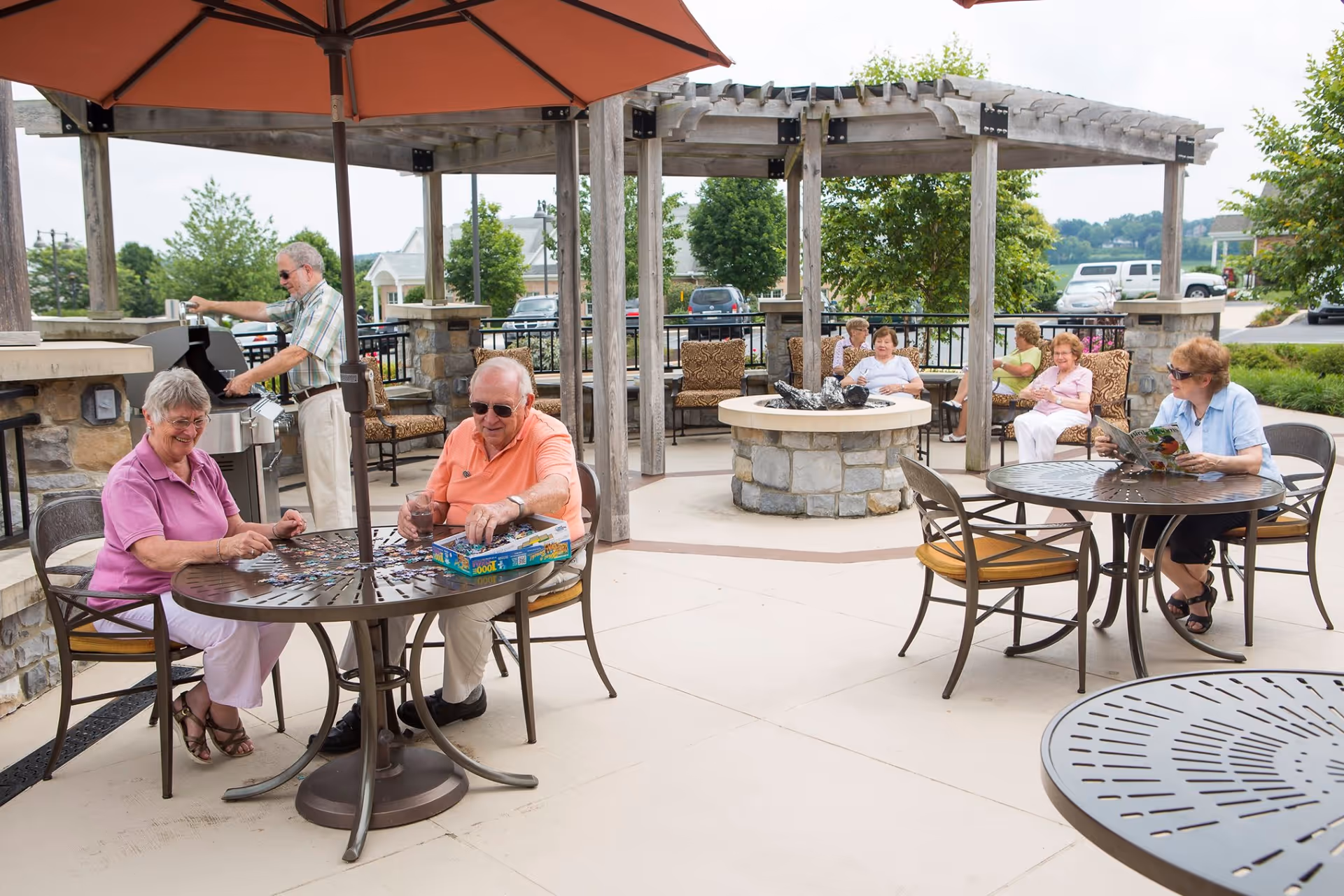A group of elderly people enjoying an outdoor patio area at Luther Acres. Two seniors are sitting at a round table under an umbrella working on a jigsaw puzzle. Another senior is grilling food on a barbecue. In the background, several seniors are seated on cushioned chairs around a stone fire pit under a wooden pergola, while one woman is reading a magazine at another table.